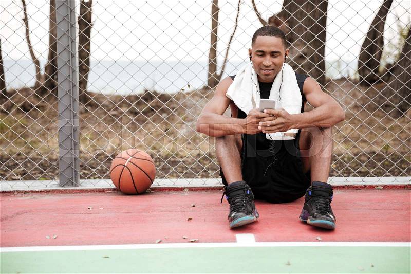 Picture of young happy african basketball player sitting in the park with towel near basketball and chatting while listen to music. Look at phone, stock photo