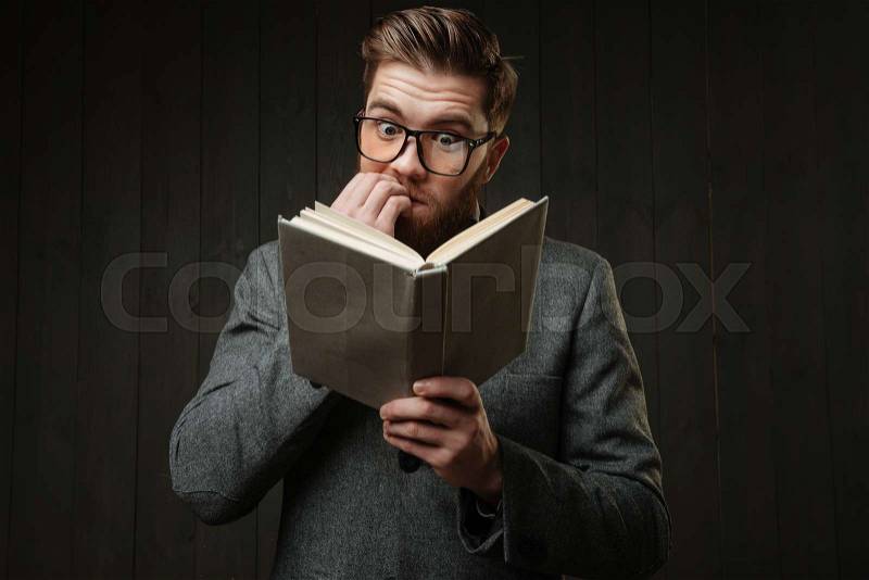 Portrait of a nervous man in eyeglasses reading book and biting nails isolated on the black wooden background, stock photo