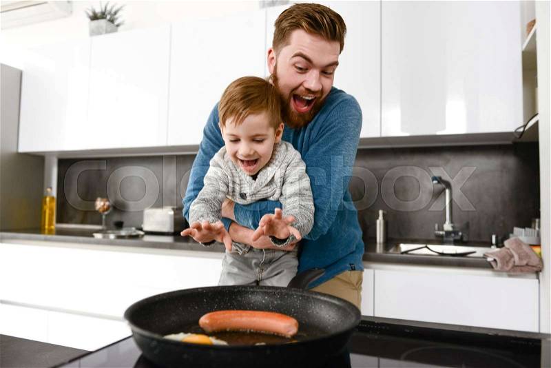 Photo of cheerful bearded father dressed in blue sweater cooking at kitchen with his little cute son, stock photo