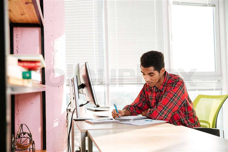 Picture of young african student writing notes in notebook while sitting near computer, stock photo
