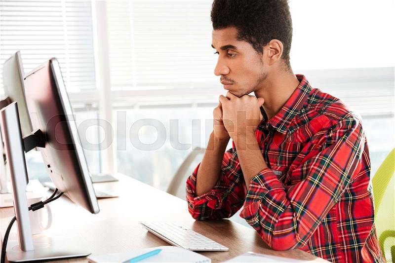 Image of handsome african guy dressed in shirt in a cage print student sitting in room while looking at computer, stock photo