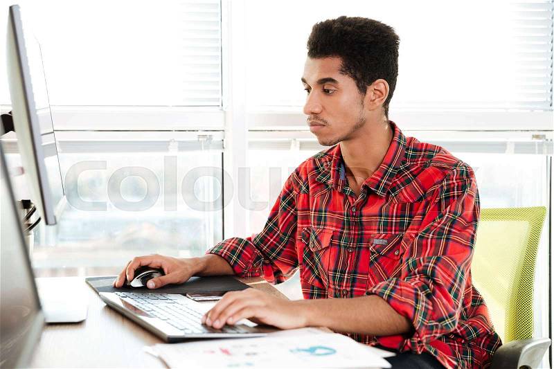 Image of concentrated handsome african student sitting in room and typing by keyboard while looking at computer, stock photo