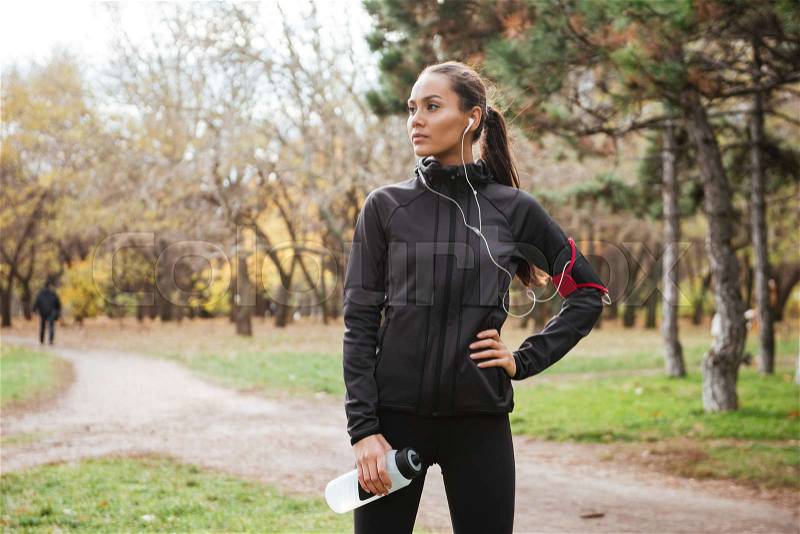 Female young runner in warm clothes looking aside in autumn park while holding bottle of water, stock photo