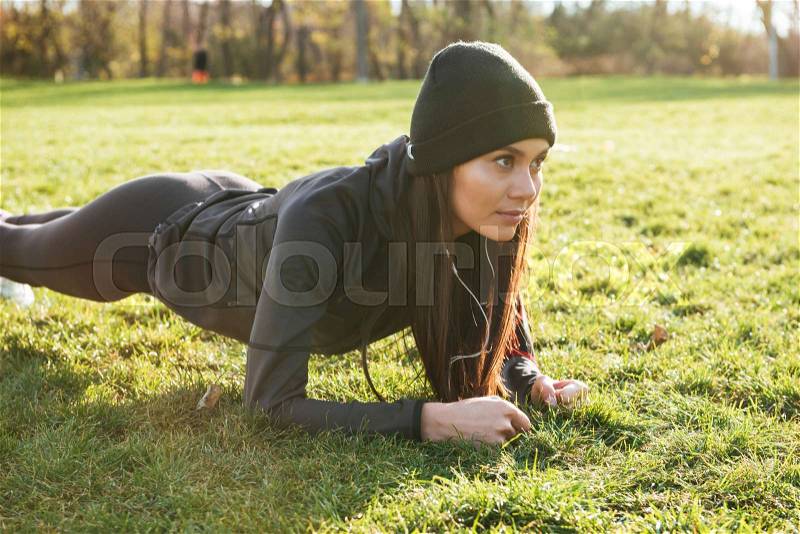 Image of young fitness lady in warm clothes and headphones in autumn park make plank, stock photo