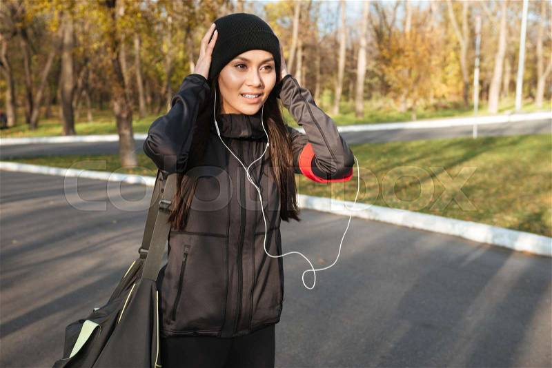 Young fitness woman in warm clothes and earphones in autumn park holding bag. Look aside, stock photo