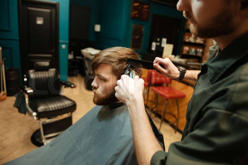 Photo of young bearded man getting haircut by hairdresser with electric razor while sitting in chair, stock photo