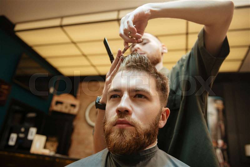Man getting haircut by hairdresser with scissors while sitting in chair. Look aside, stock photo