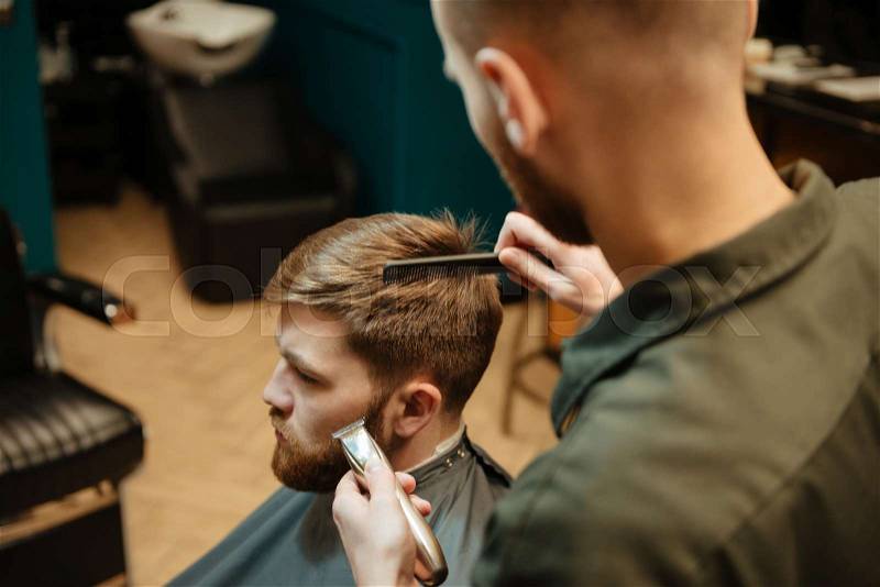 Image of young handsome man getting haircut by hairdresser with razor while sitting in chair in barbershop. Look aside, stock photo