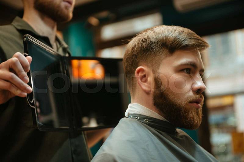 Photo of young handsome man getting haircut by hairdresser while sitting in chair. Look at mirror, stock photo