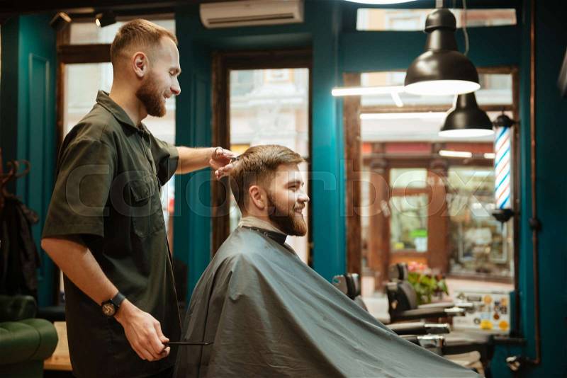 Cheerful man getting haircut by hairdresser with scissors while sitting in chair. Look aside, stock photo