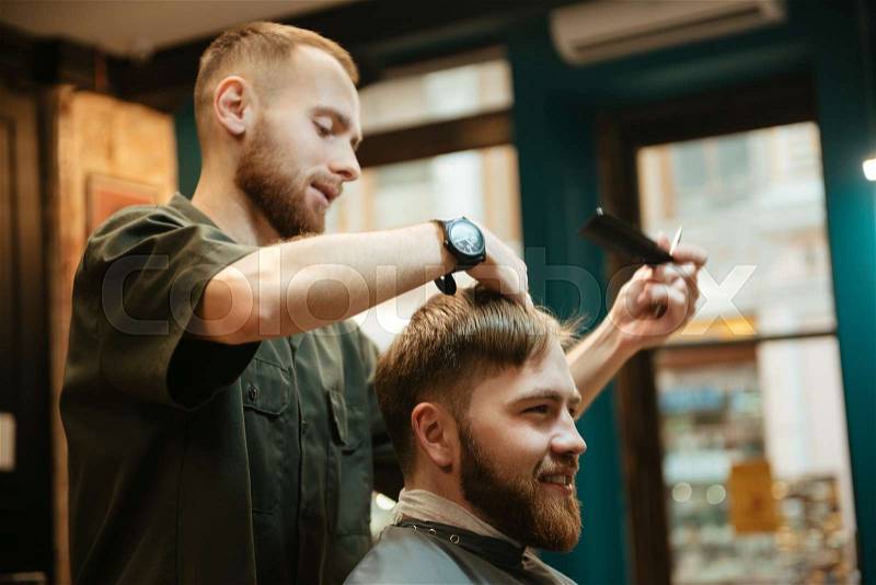 Photo of happy man getting haircut by hairdresser with scissors while sitting in chair. Look aside, stock photo