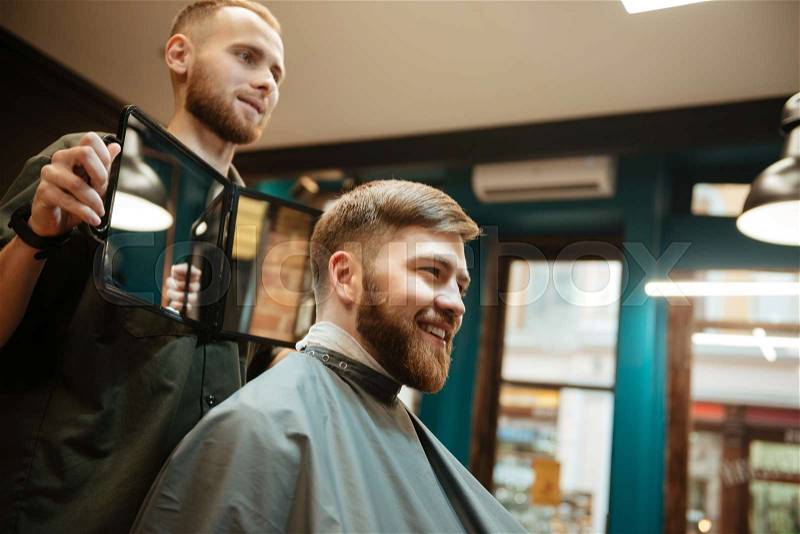 Young handsome man getting haircut by hairdresser while sitting in chair. Look at mirror, stock photo