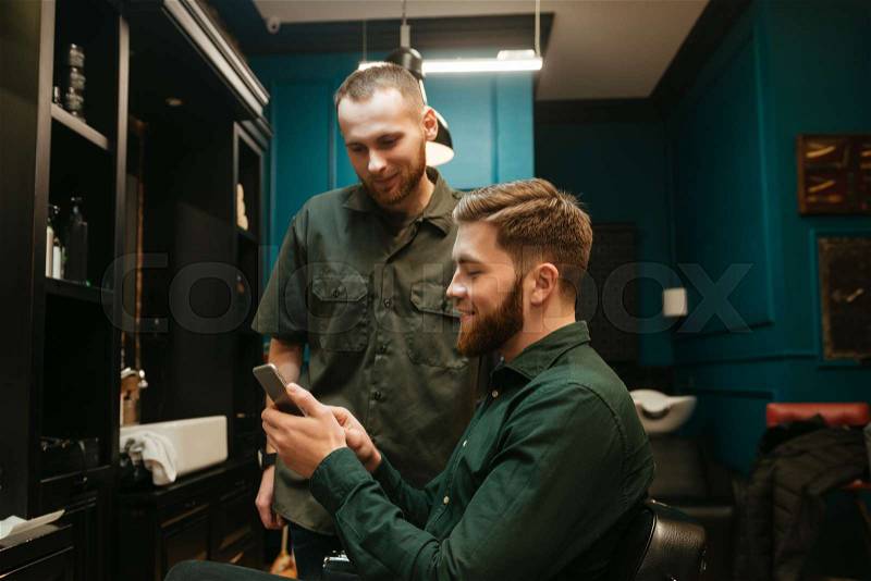 Happy young hipster man getting haircut by hairdresser while sitting in chair. Holding phone, stock photo