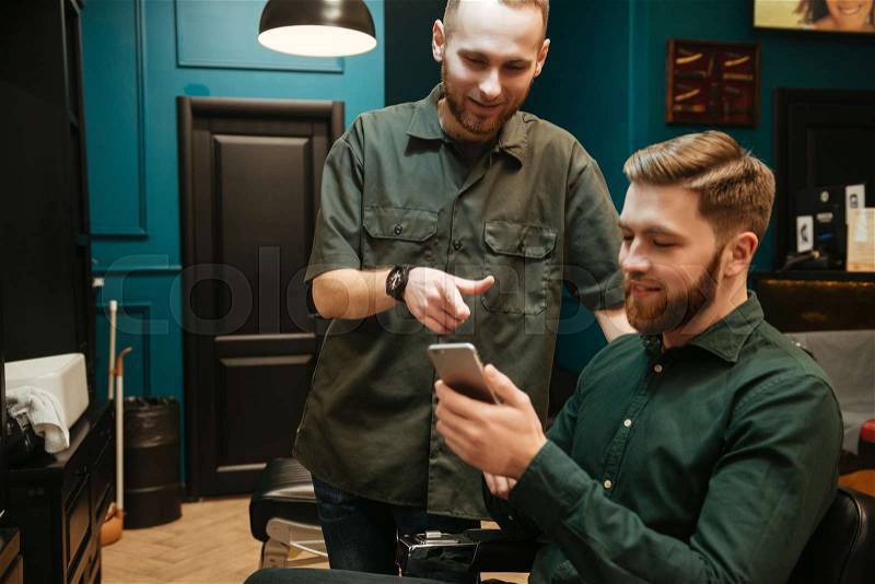 Cheerful young man getting haircut by hairdresser while sitting in chair. Holding phone, stock photo