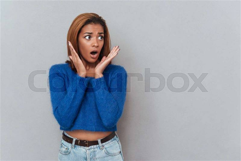 Surprised young african woman in sweater looking away and holding her hands near the face. Isolated gray background, stock photo
