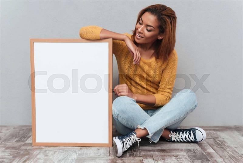Young smiling african woman in yellow sweater and jeans sitting on the wooden floor and looking at the blank board. Isolated gray background, stock photo