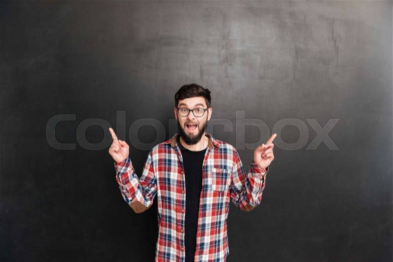 Happy bearded young man in glasses pointing away with both hands over grey background, stock photo