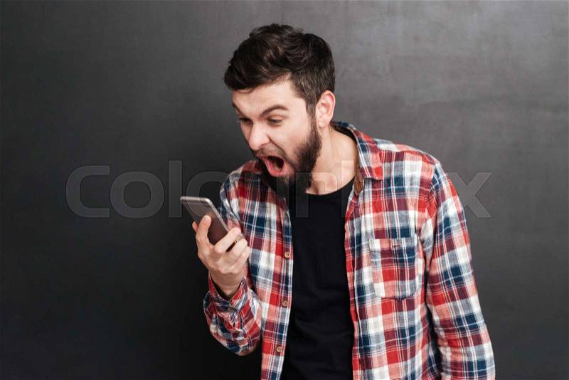 Mad irritated bearded young man in plaid shirt holding and shouting on cell phone over grey background, stock photo
