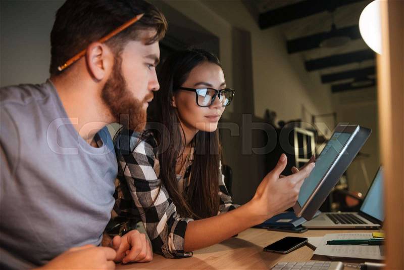 Concentrated colleagues working late at night in their office with tablet computer. Woman talking to man. Man looking at tablet computer, stock photo