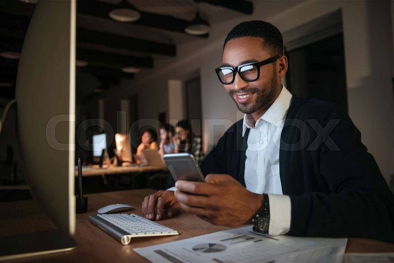 Happy dark skinned businessman working late at night in office with computer. Chatting by phone, stock photo