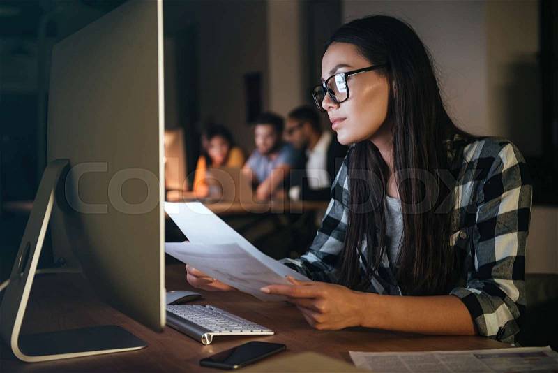 Concentrated businesswoman wearing glasses working late at night in office with computer. Looking at computer while holding documents, stock photo