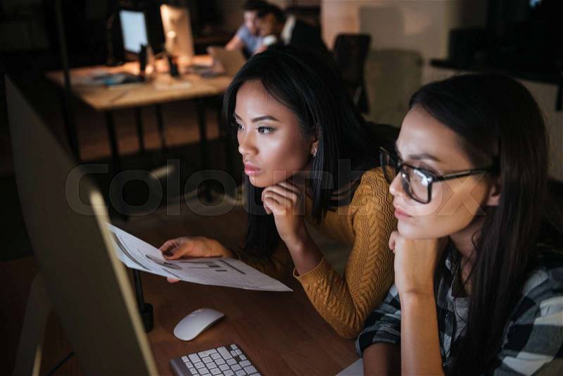 Two pretty businesswomen working late at night in office with computer. Looking at computer, stock photo