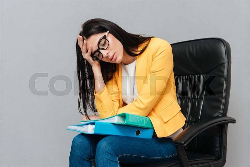 Tired woman wearing eyeglasses and dressed in yellow jacket holding folders while sitting on office chair over grey background. Eyes closed, stock photo