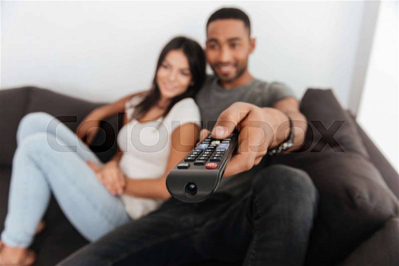 Picture of happy young couple hugging and watching TV on sofa at home. Focus on hand with remote control, stock photo