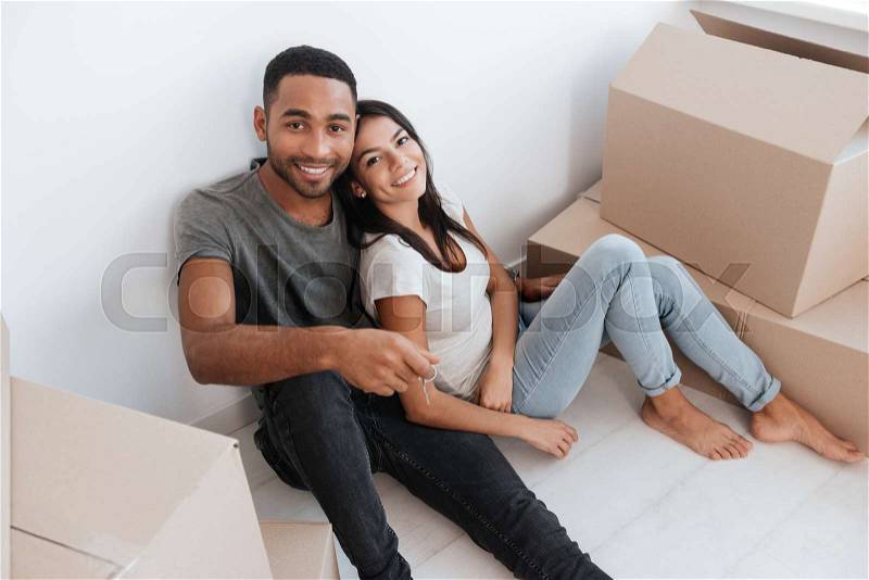 Joyful couple sitting with unpacked boxes. Man holding key. Looking at camera, stock photo