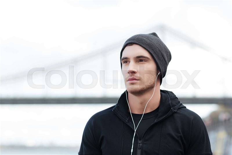 Handsome young man in hat listening to music with earphones outdoors, stock photo