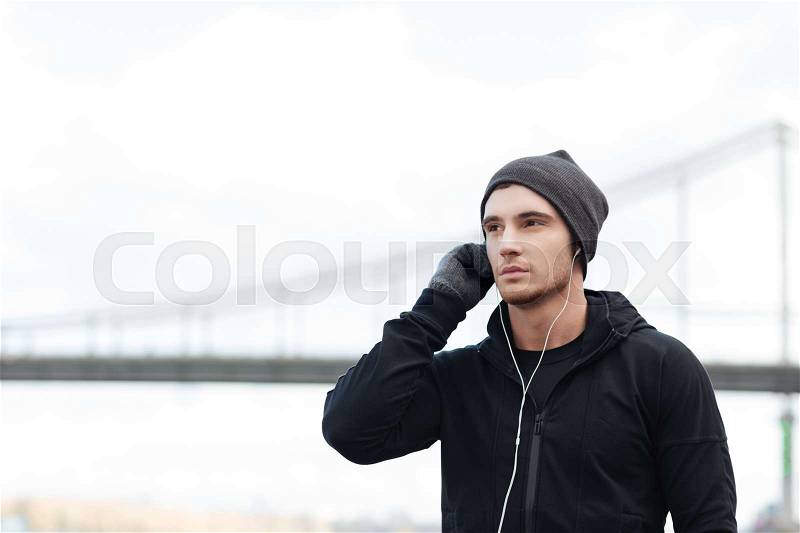 Serious young man in hat and gloves listening to music with earphones outdoors, stock photo