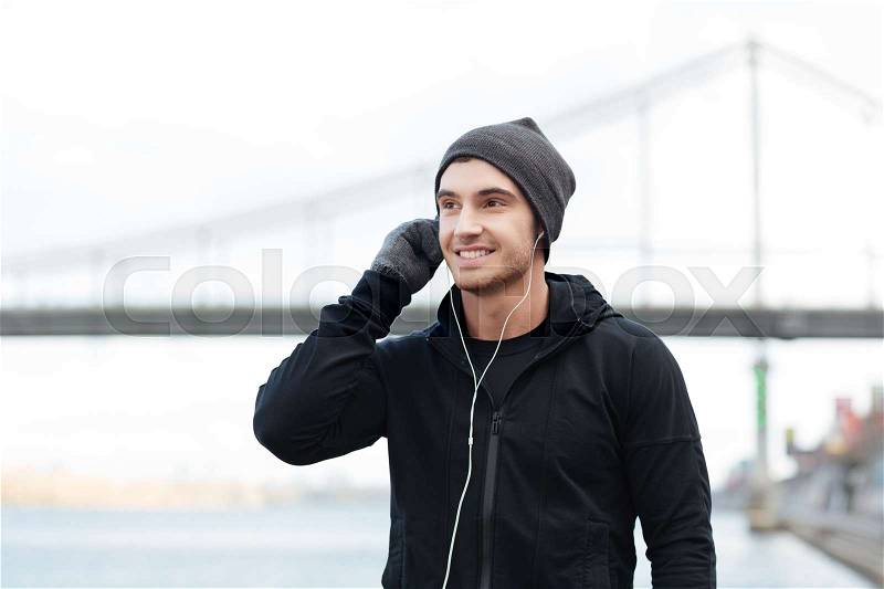 Happy young man in hat and gloves listening to music with earphones outdoors, stock photo