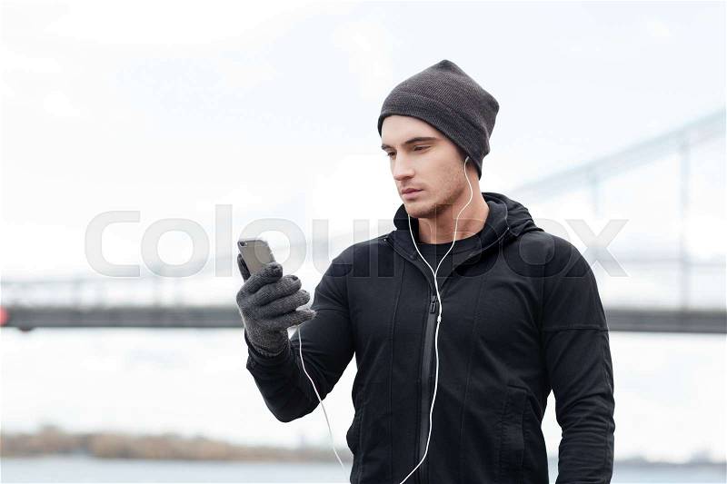 Concentrated young man athlete standing and listening to music with mobile phone and earphones outdoors, stock photo