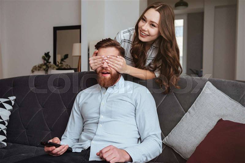 Photo of cheerful woman covering man\'s eyes while he sitting at sofa, stock photo
