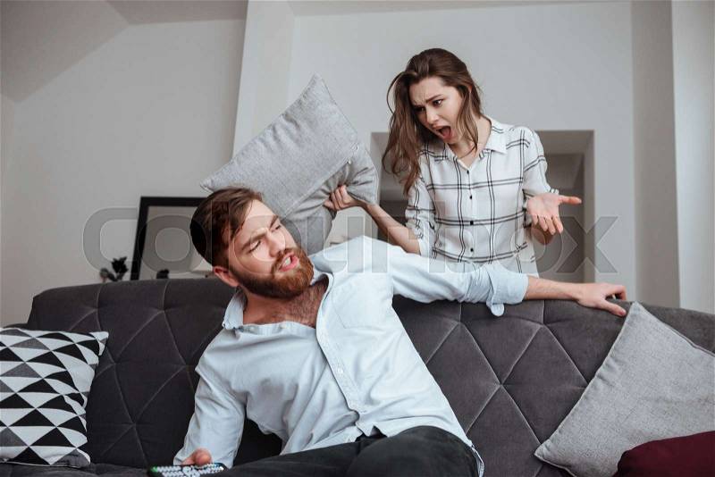 Image of angry woman screaming to man while he sitting at sofa. Look at each other, stock photo