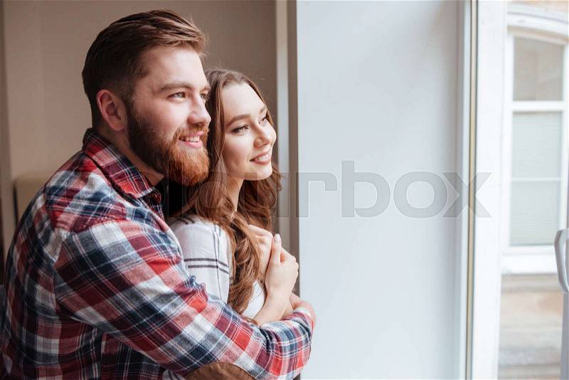 Happy boyfriend and girlfriend standing near window while hugging. Look at window, stock photo