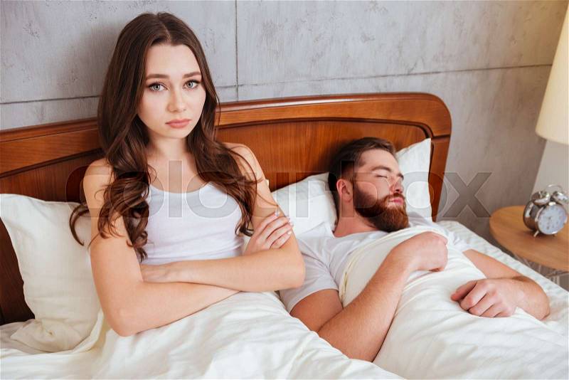 Photo of young confused woman with arms crossed looking at camera while her boyfriend sleeping, stock photo