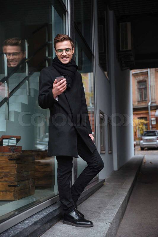 Vertical image of smiling business man in glasses with phone near the building. man looking at phone, stock photo