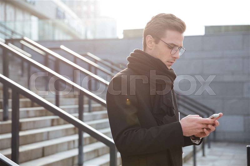 Side view of Business man in glasses and warm clothes writing message on phone and standing on the stairs, stock photo