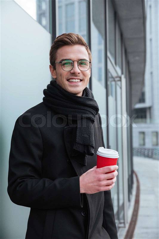 Vertical image of Business man in glasses and warm clothes standing sideways and drinking coffee outdoors, stock photo