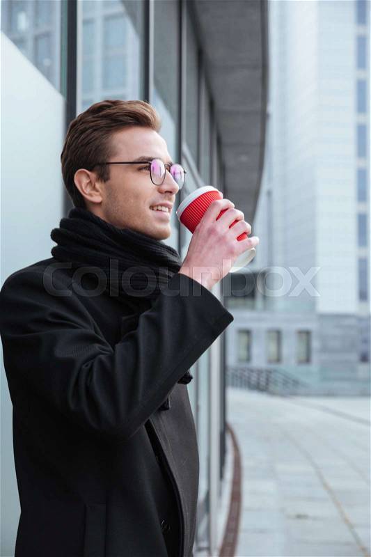 Vertical image of young business man in glasses and warm clothes standing sideways and drinking coffee outdoors, stock photo