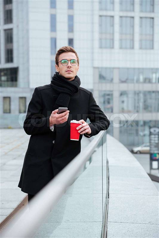 Vertical image of business man in glasses and warm clothes with coffee and phone outdoors, stock photo