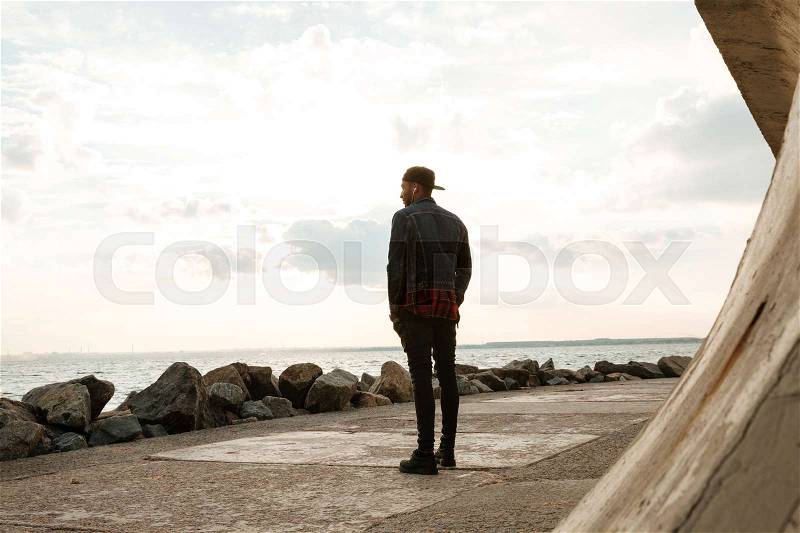 Back view of african young man wearing cap walking on the beach while listening music with earphones. Look at sea, stock photo
