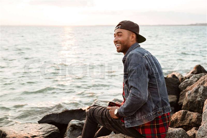 Picture of african happy man wearing cap walking on the beach. Looking aside, stock photo