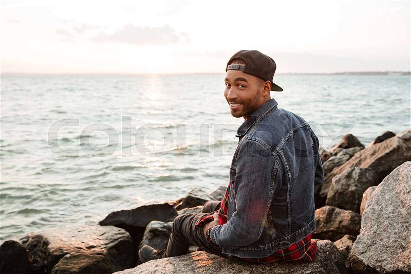 Photo of african happy man wearing cap walking on the beach. Looking at camera, stock photo