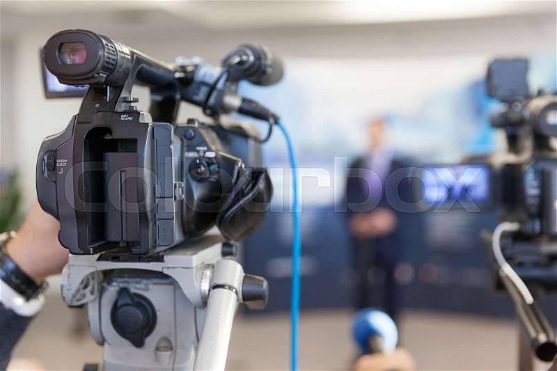 Video camera in focus, blurred spokesperson in background. Corporate news conference, stock photo
