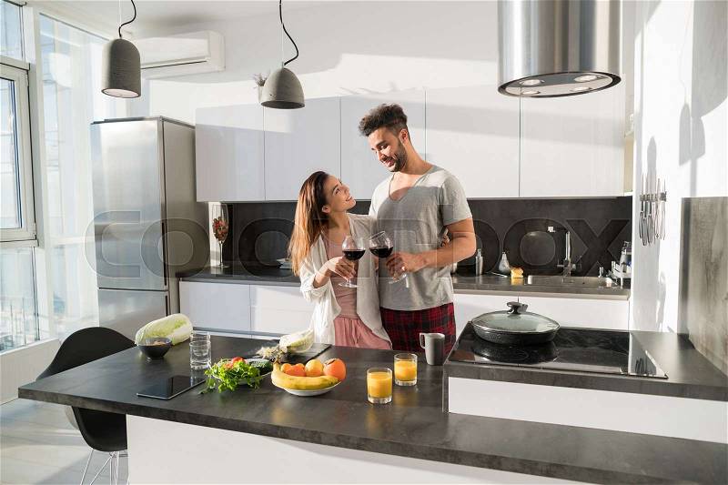 Young Couple Embrace In Kitchen, Hispanic Man And Asian Woman Hug Drink Wine Modern Apartment Interior, stock photo