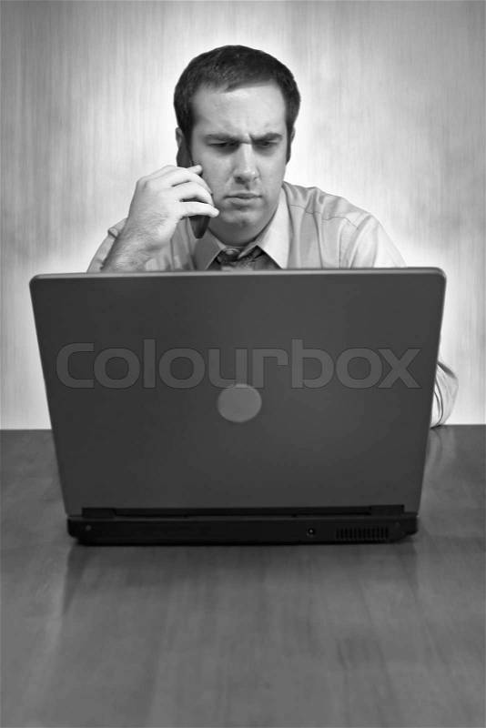 A man working from home with his cell phone and laptop in black and white He has an upset or serious look on his face, stock photo