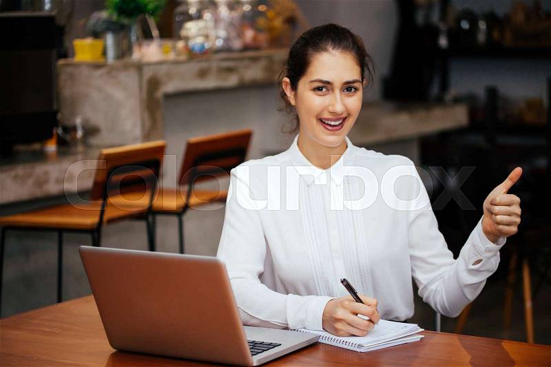 Young Caucasian woman showing thumbs up with laptop and notebook in the modern interior office - Gesture and thumbs up sign, stock photo