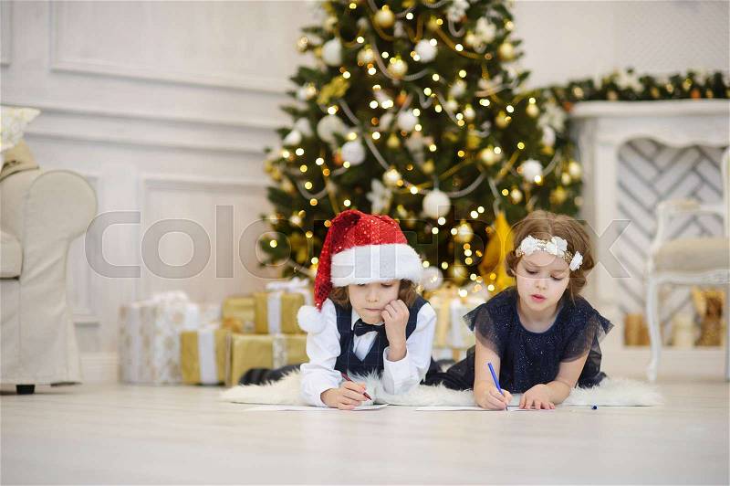 Christmas Eve. Children write letters to Santa Claus. Boy and girl of younger school age lie on a floor near the decorated Christmas tree. Children have thoughtful faces. House is festively decorated, stock photo
